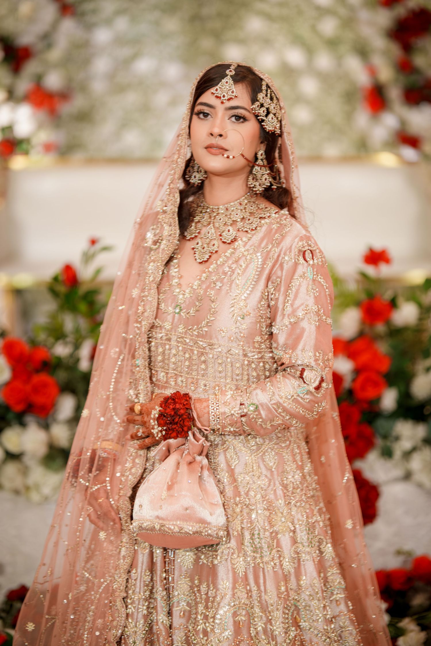 Bride in ornate peach sequined lehenga and veil poses against backdrop of red and white roses.