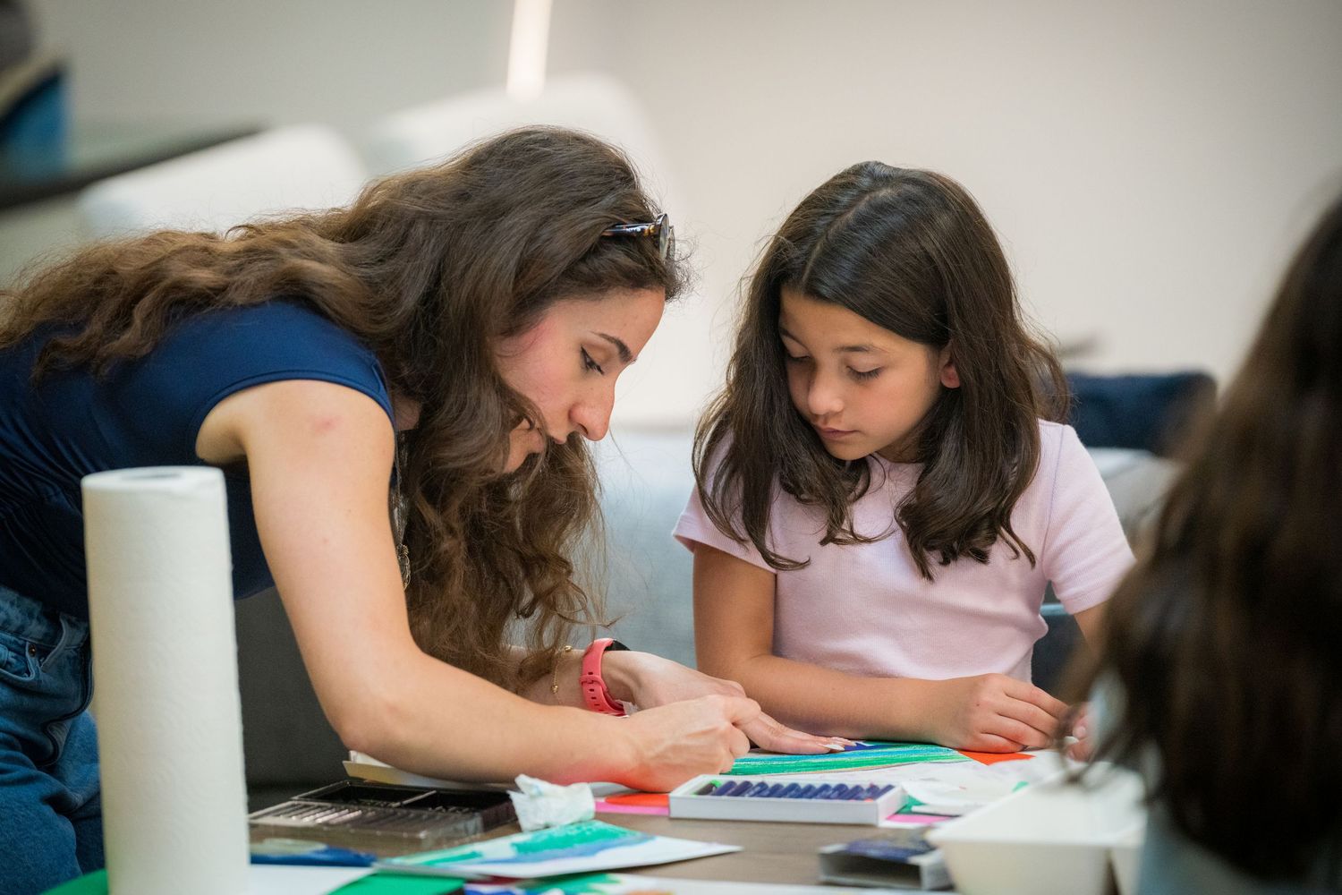 A teacher helps a young girl with a colorful art project at a table in a classroom.
