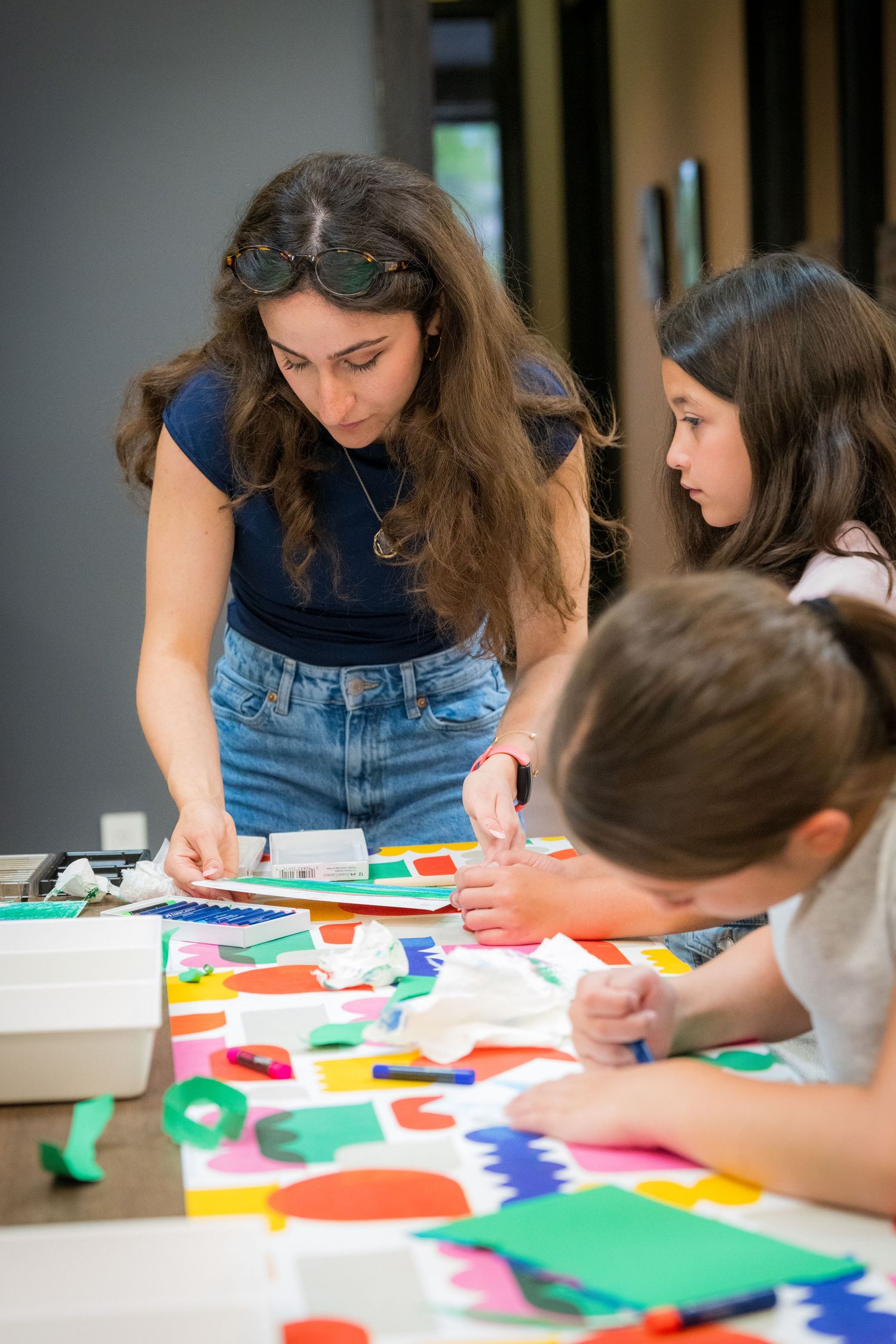 A teacher guides two children working on a colorful paper collage art project.