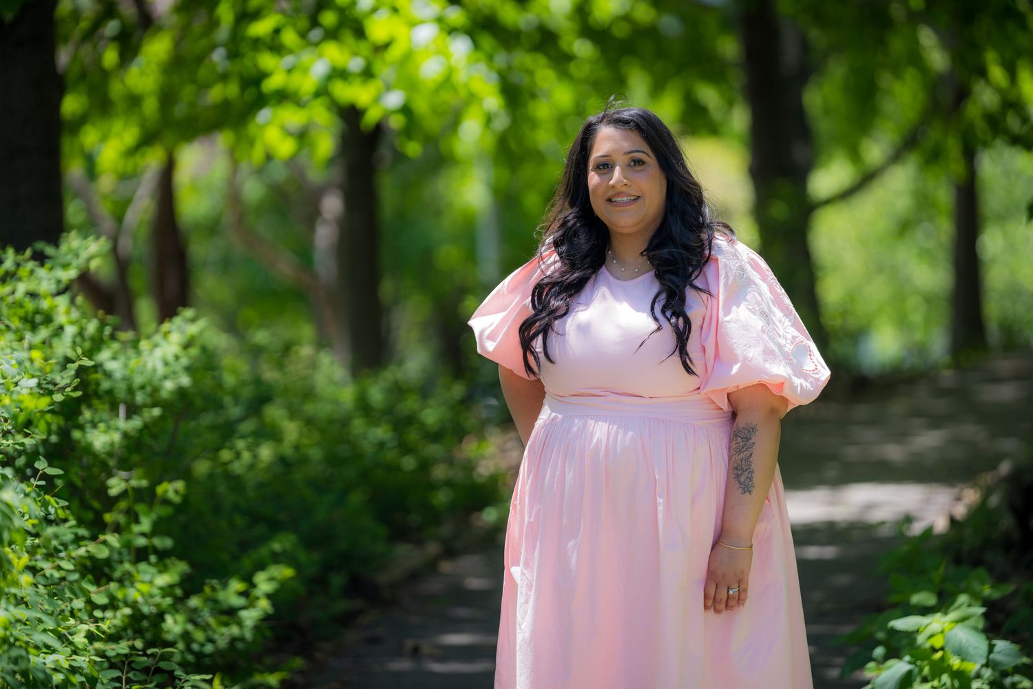 Pregnant woman in pink maternity dress smiling on a tree-lined garden path on a sunny day.