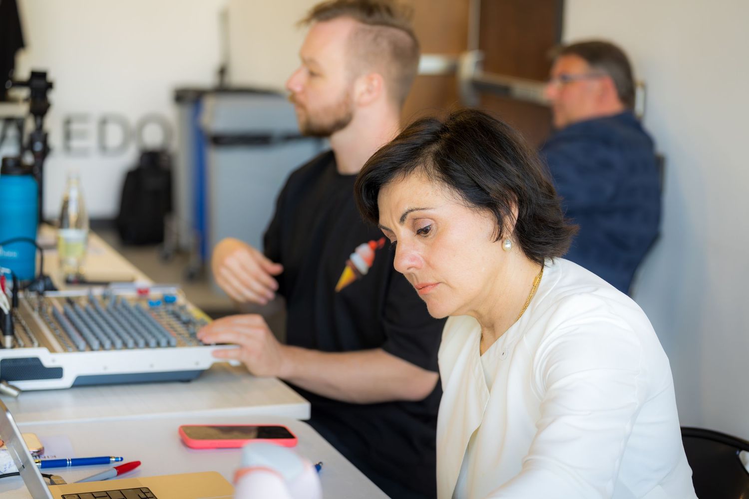 Woman with dark hair works at desk with laptop alongside colleagues in modern office.