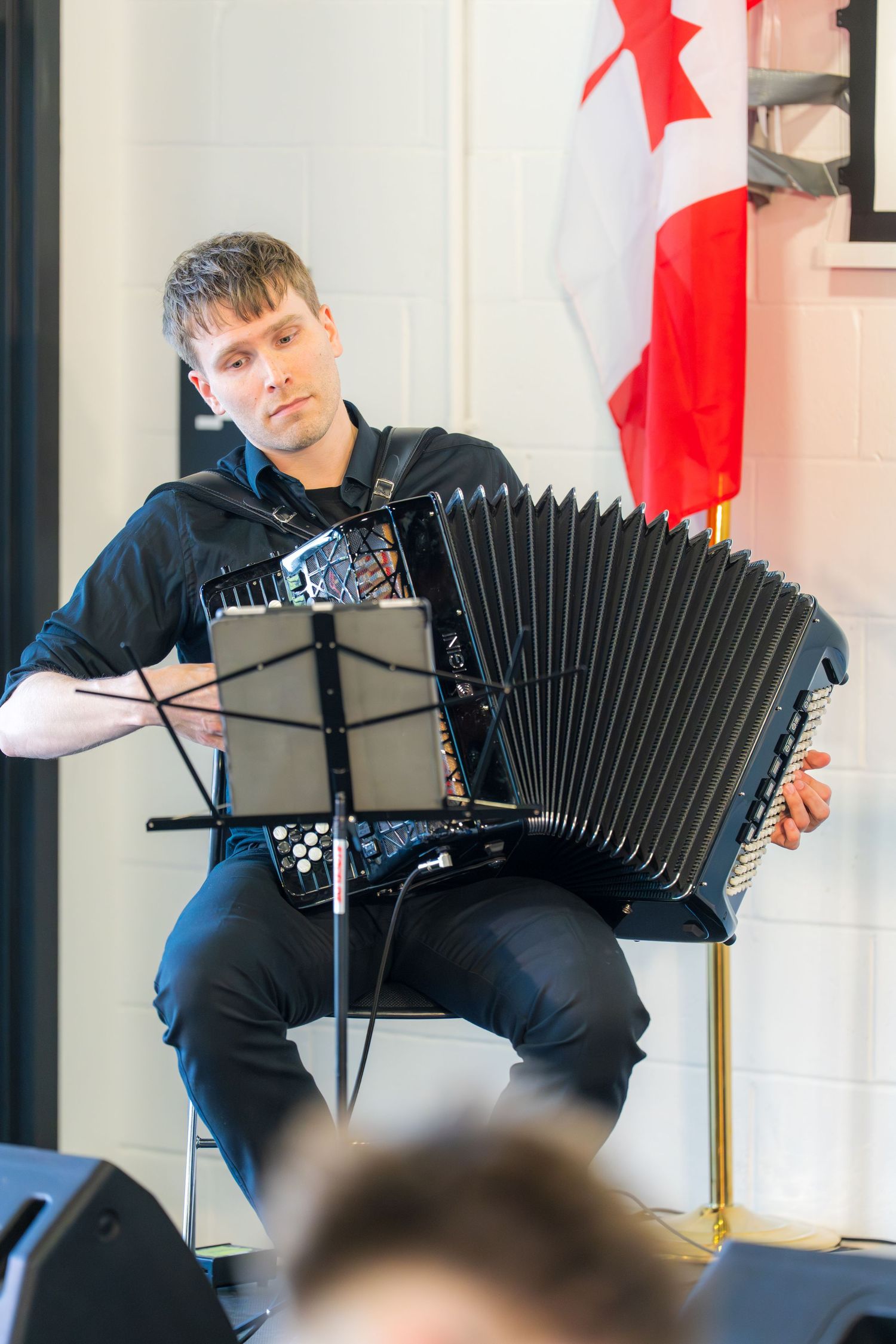 Musician playing black accordion seated on stage near a Canadian flag during a live Neapolitan Ice Cream event.