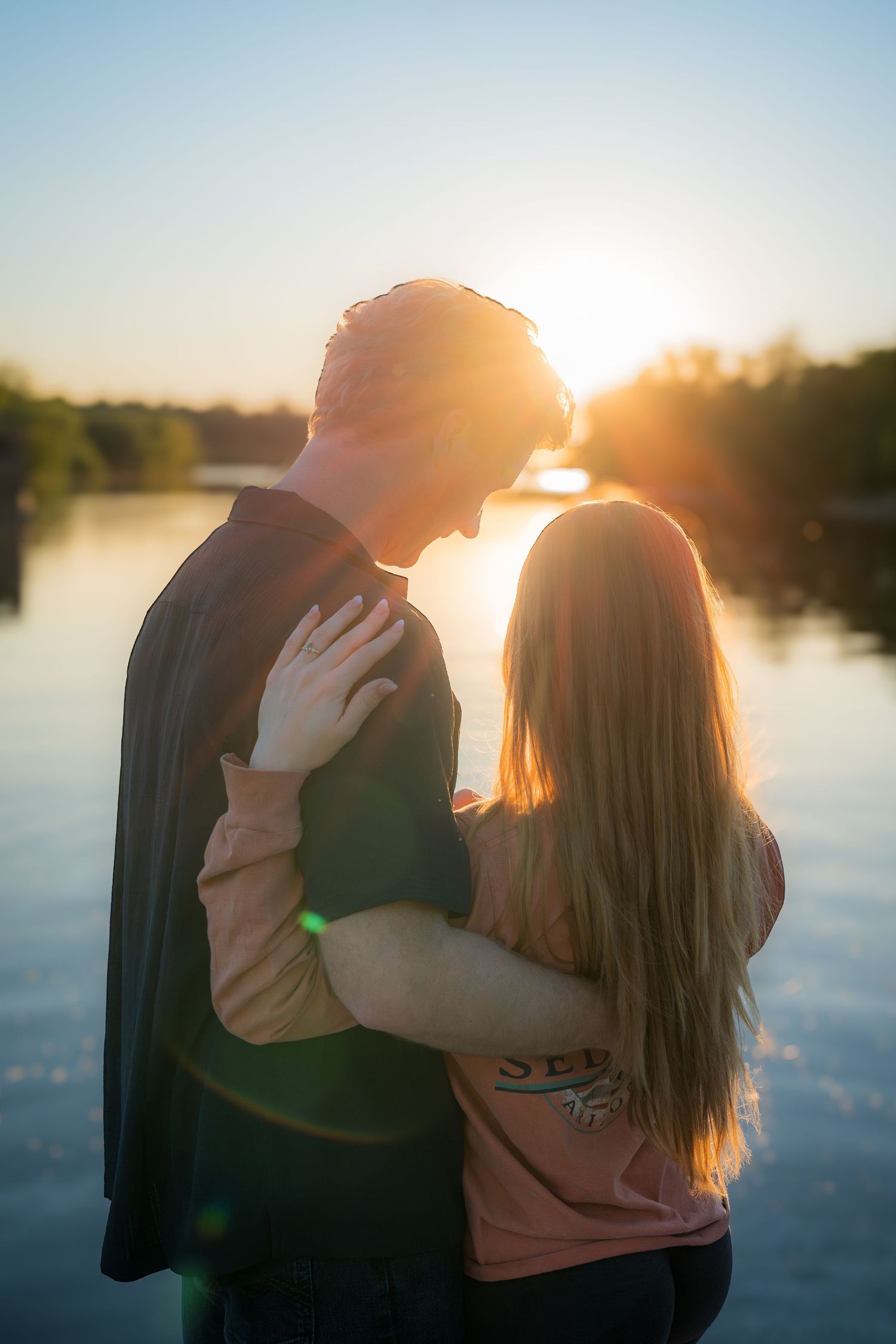 Romantic couple embracing at sunset by a lake, silhouetted against golden sunlight with lens flare effect.
