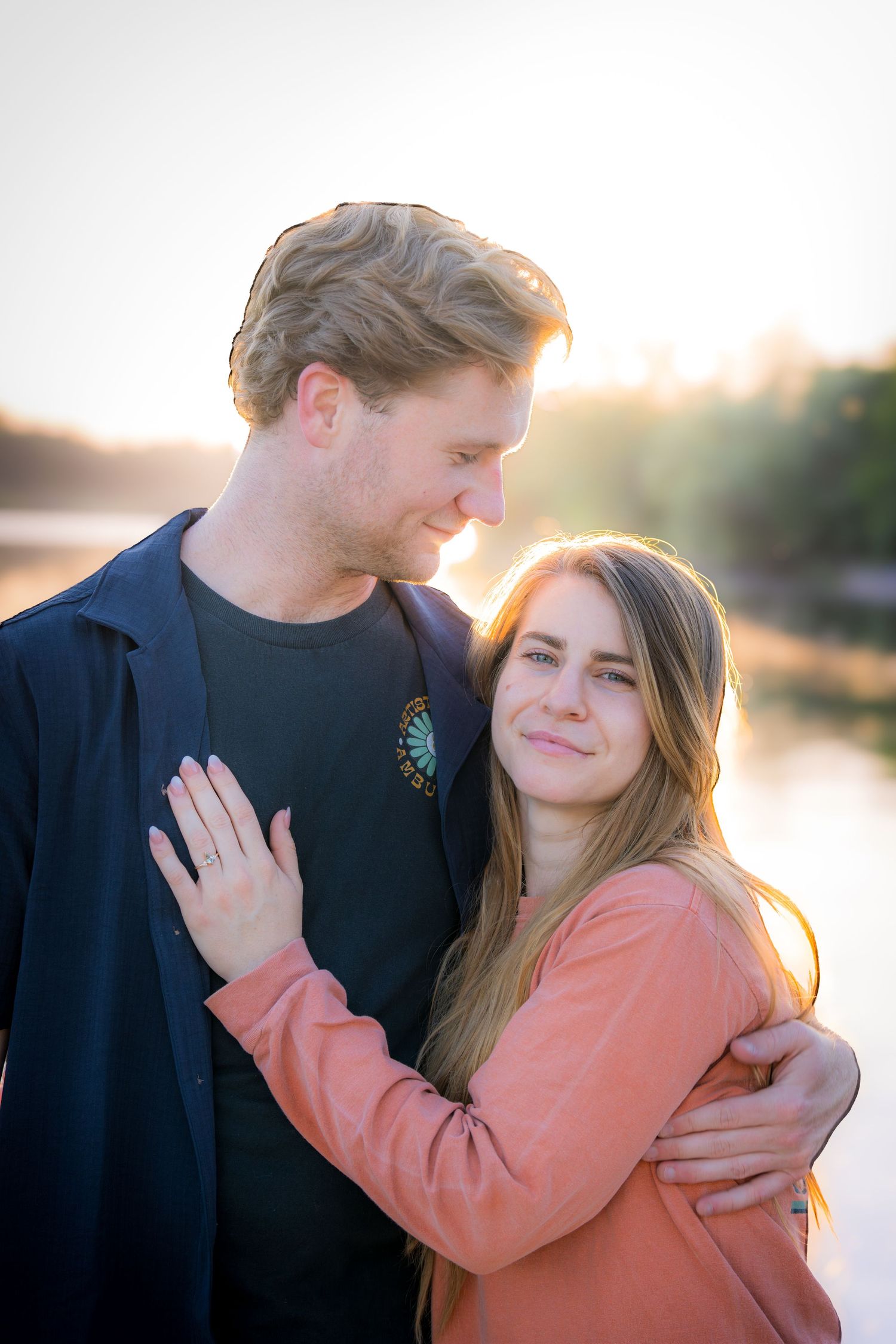 Happy engaged couple embracing near a river at golden hour with warm sunset light behind them.