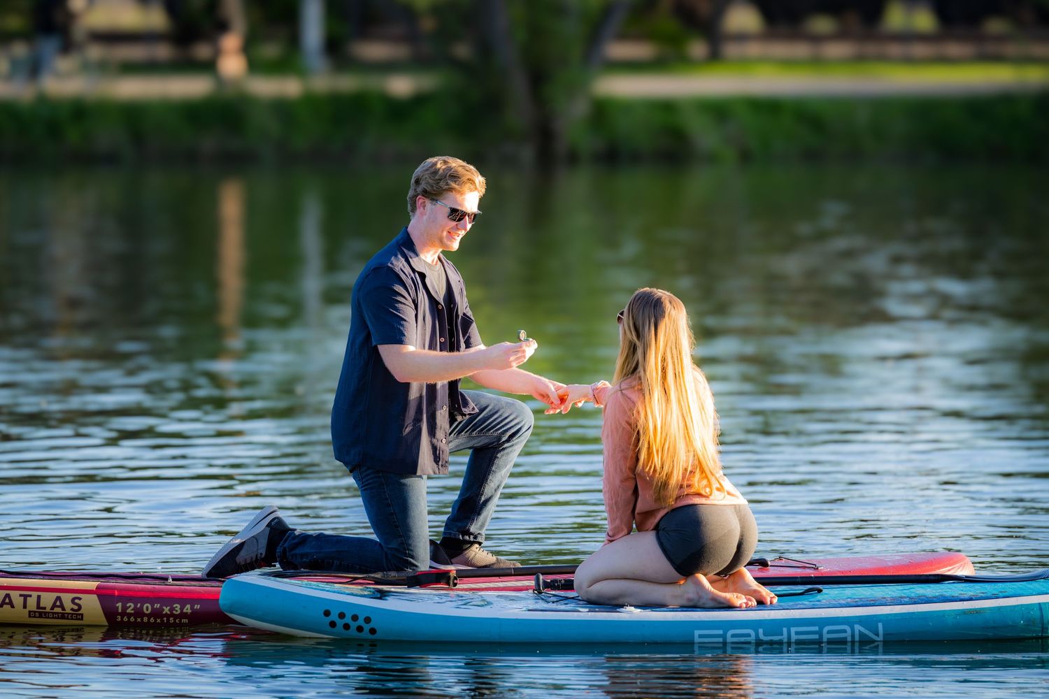Man proposing on a paddleboard to his girlfriend on a sunlit lake.