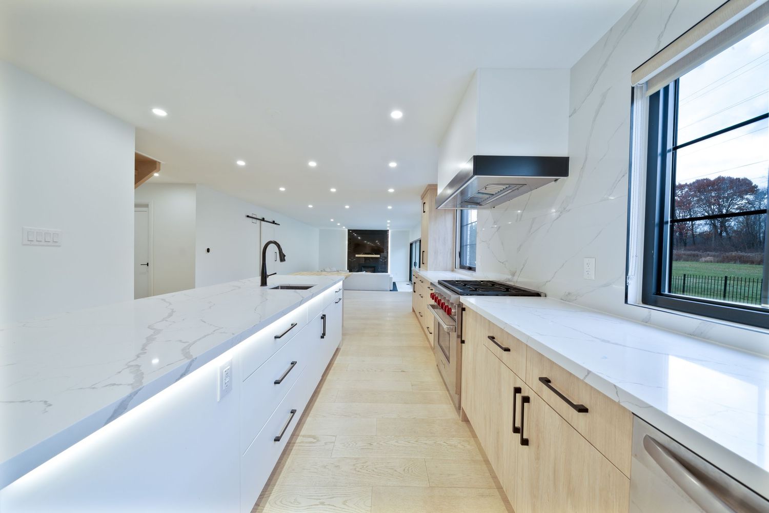 Modern galley kitchen with white cabinets, marble countertops, stainless appliances and large window overlooking green space.