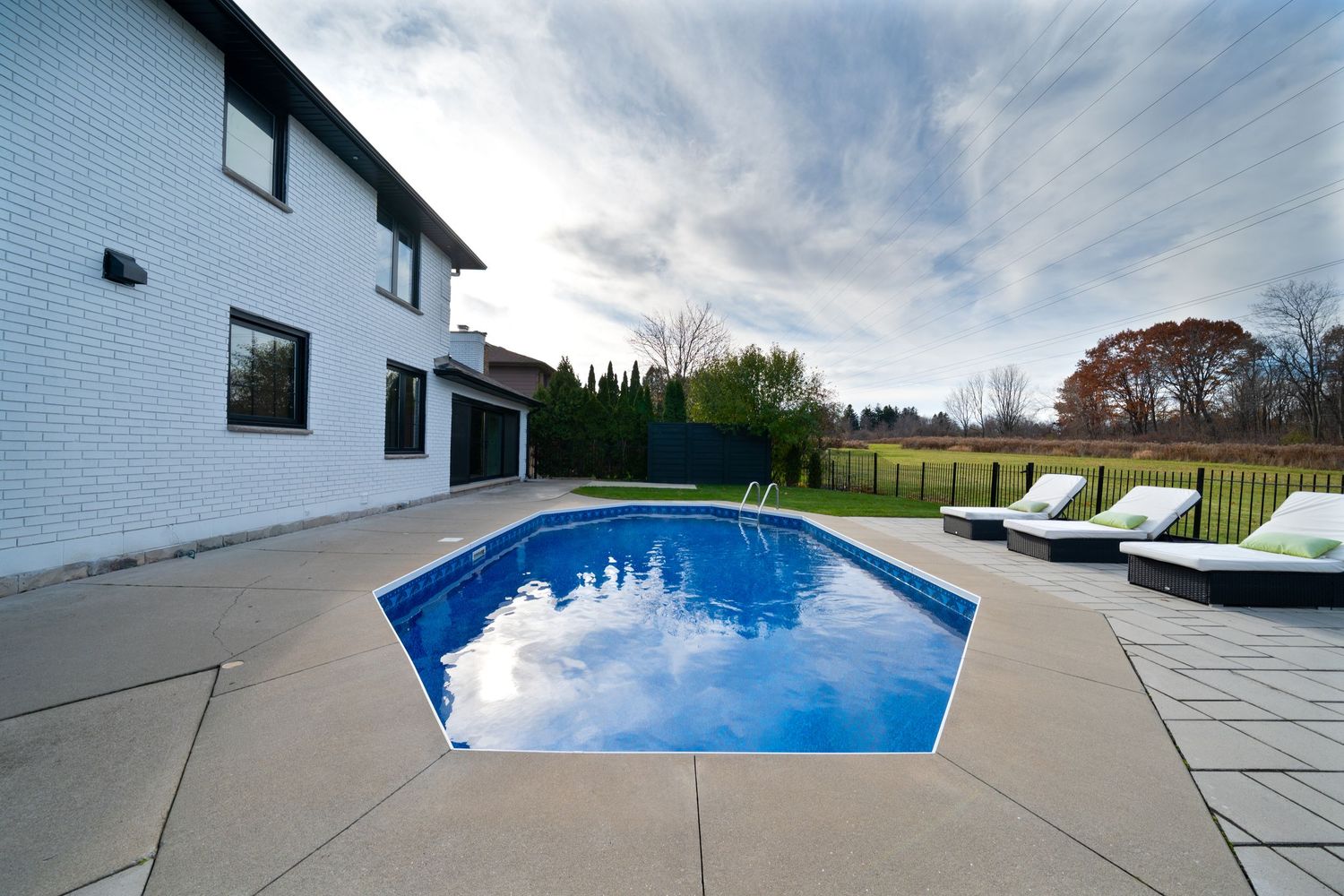 Panoramic view of a luxurious backyard pool with concrete deck and white house under cloudy blue skies.