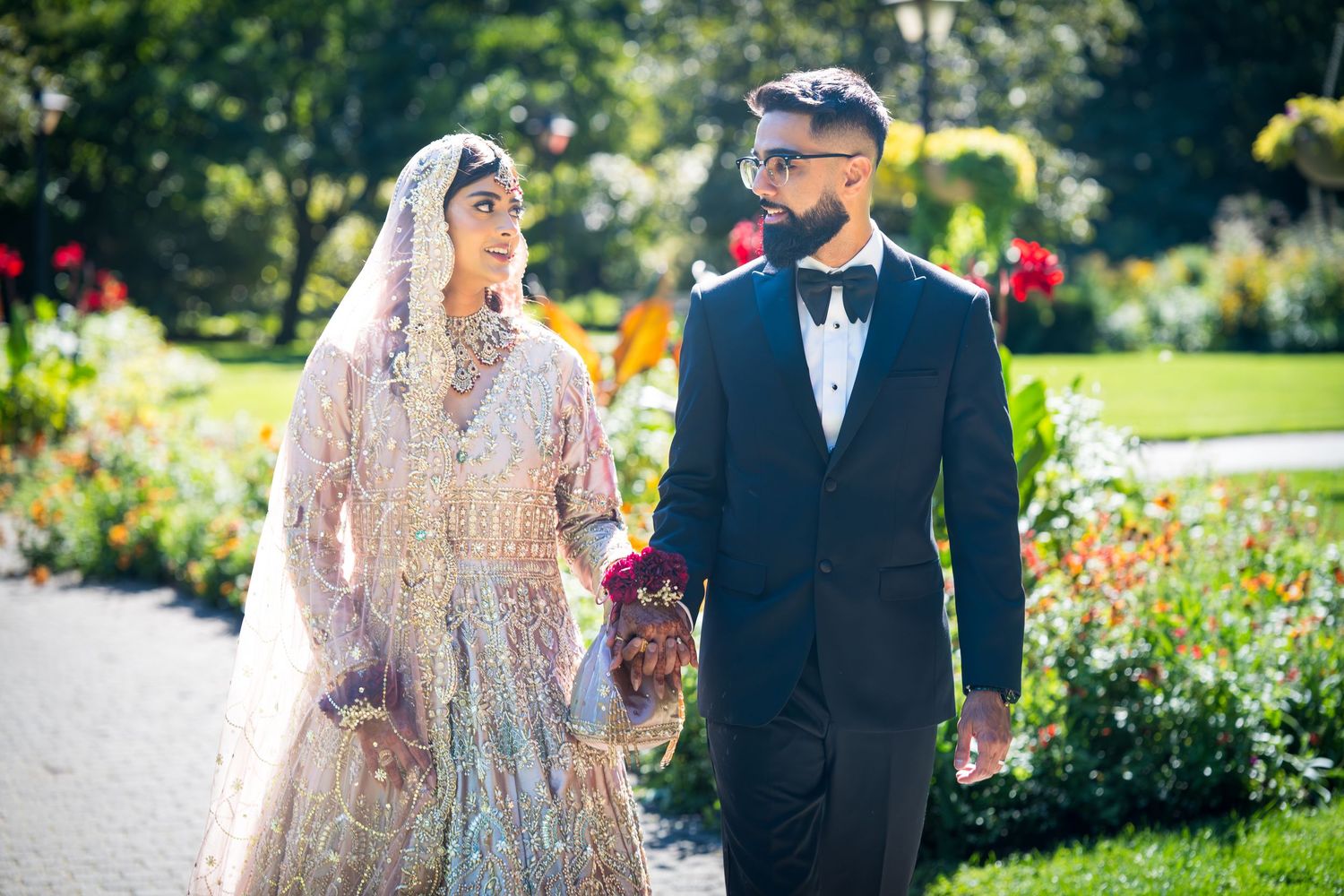 Newlyweds in elegant wedding attire stand together in a sunny garden with blooming flowers.