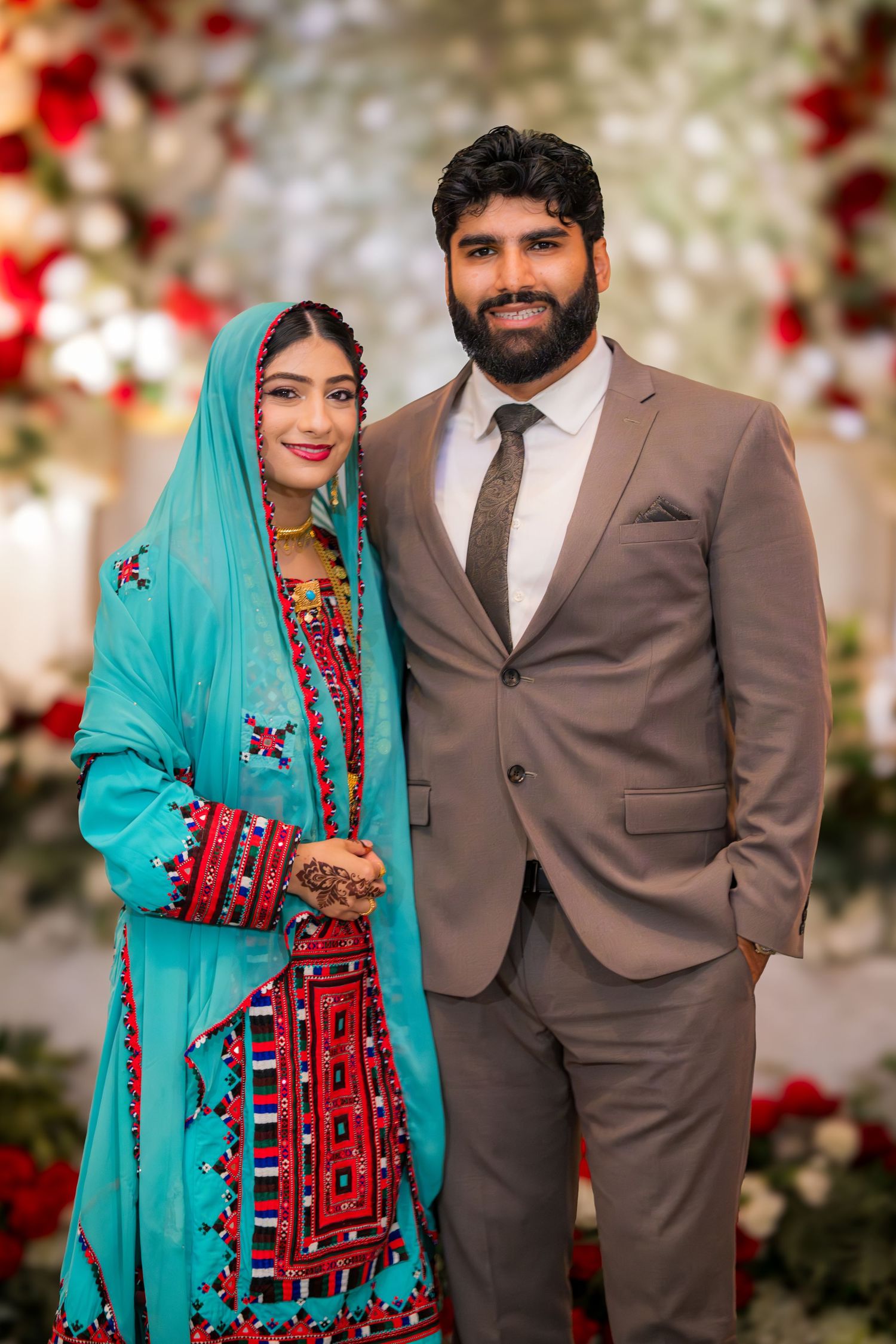 Wedding couple in traditional attire posing against floral backdrop with red roses.