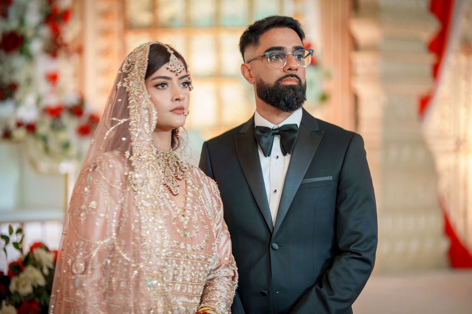 Bride in blush pink lehenga and groom in black tuxedo at an elegantly decorated wedding venue.