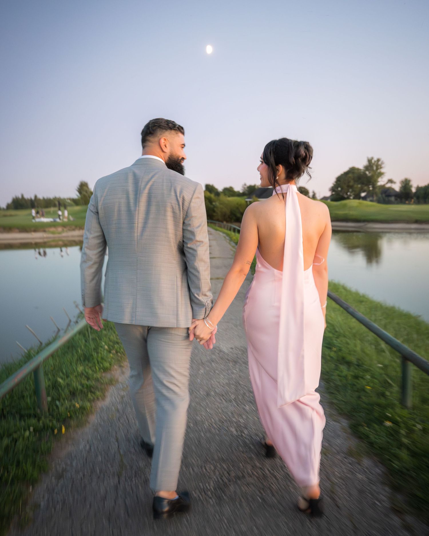 Couple in formal attire walks hand in hand along lakeside path at dusk.