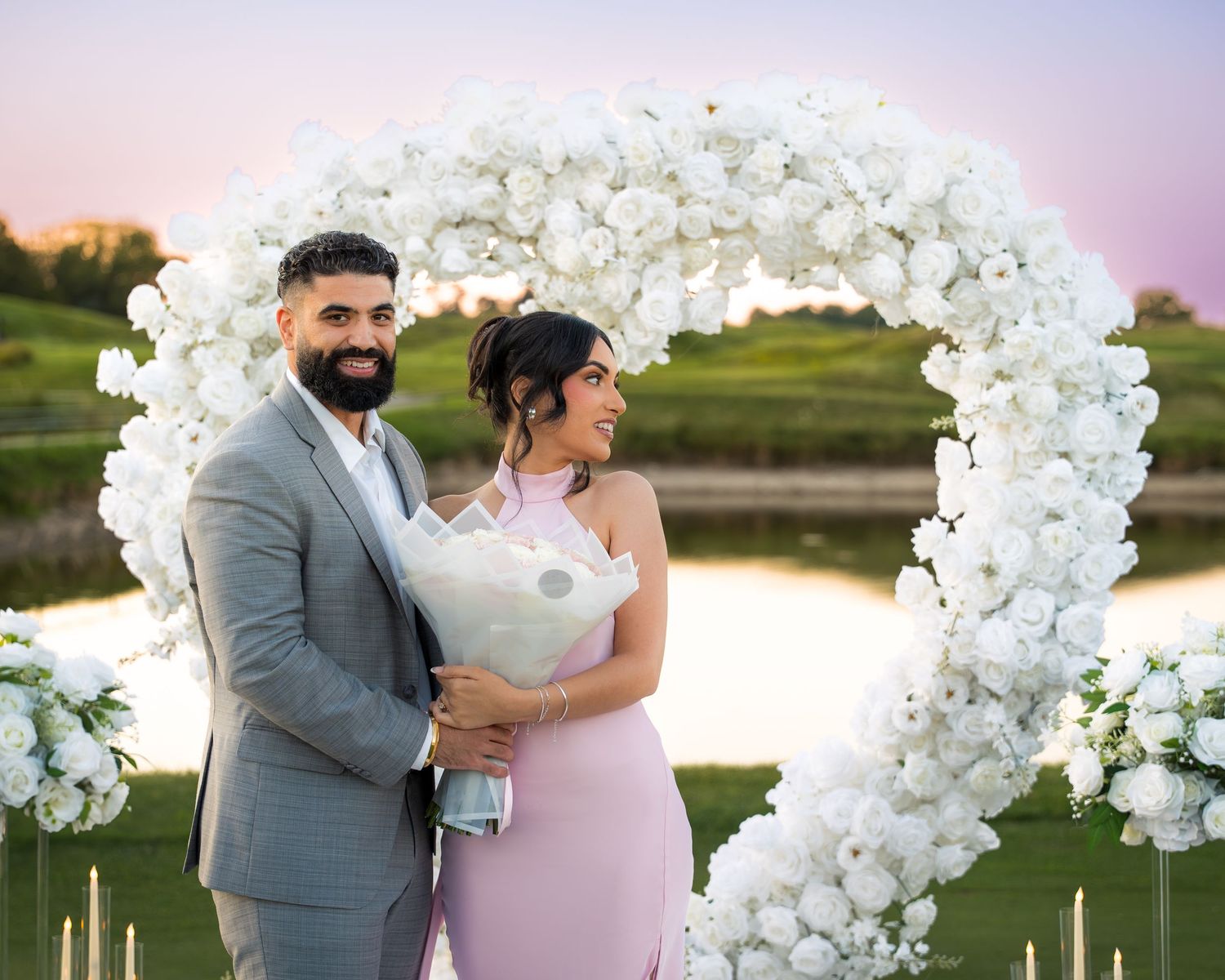 Beautiful sunset proposal moment surrounded by white floral arches and glowing candles.