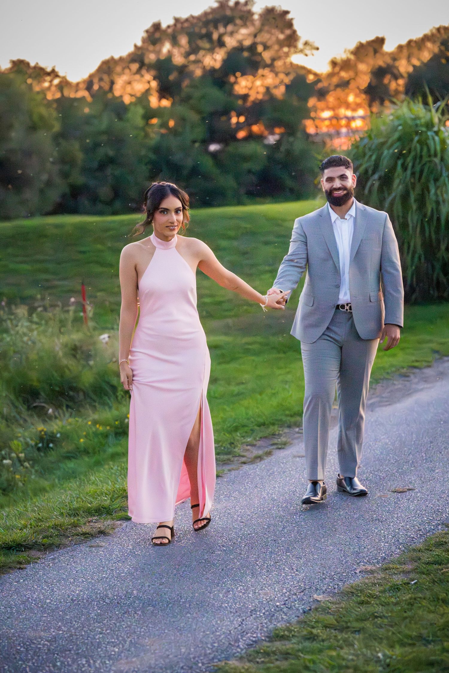 A couple in pink and grey formal attire walks together on a gravel path during golden hour sunset.