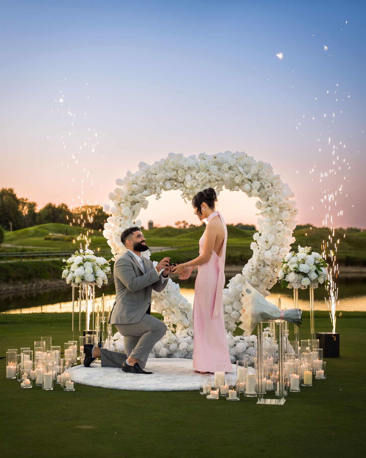 Elegant outdoor proposal scene with white floral arch and sparkling fireworks at dusk.