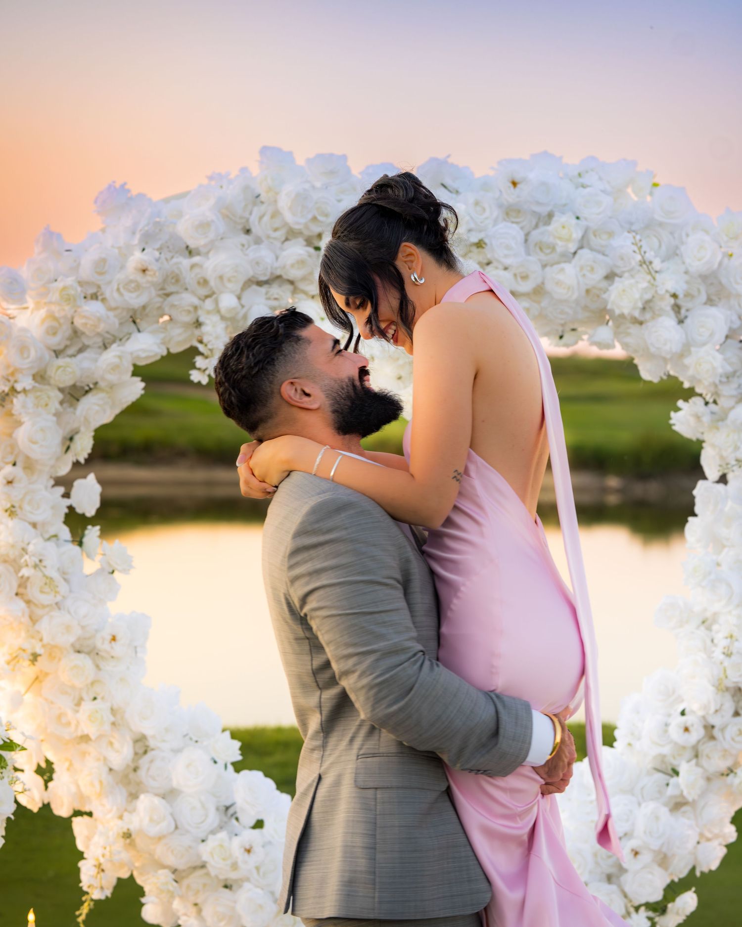 Joyful embrace after proposal under white floral arch at sunset.