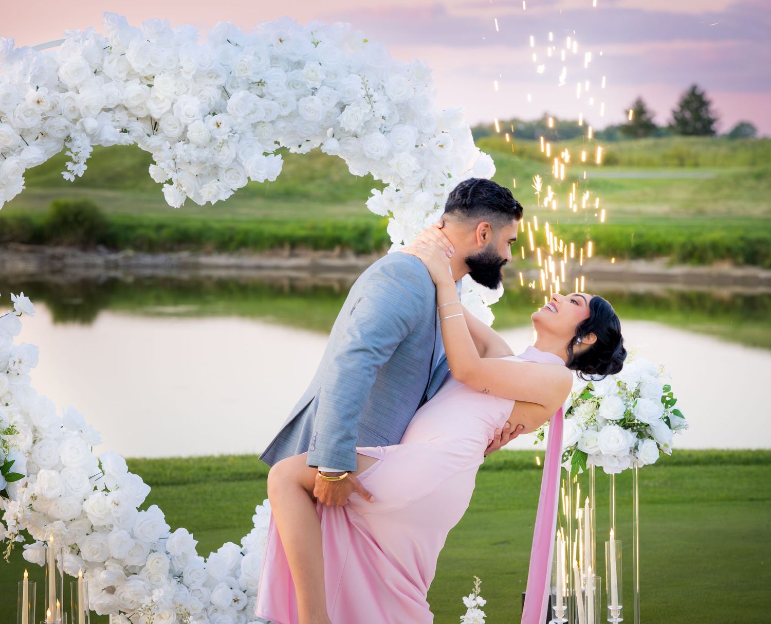A couple shares an intimate moment outdoors with white floral arch and lit candles in the background.