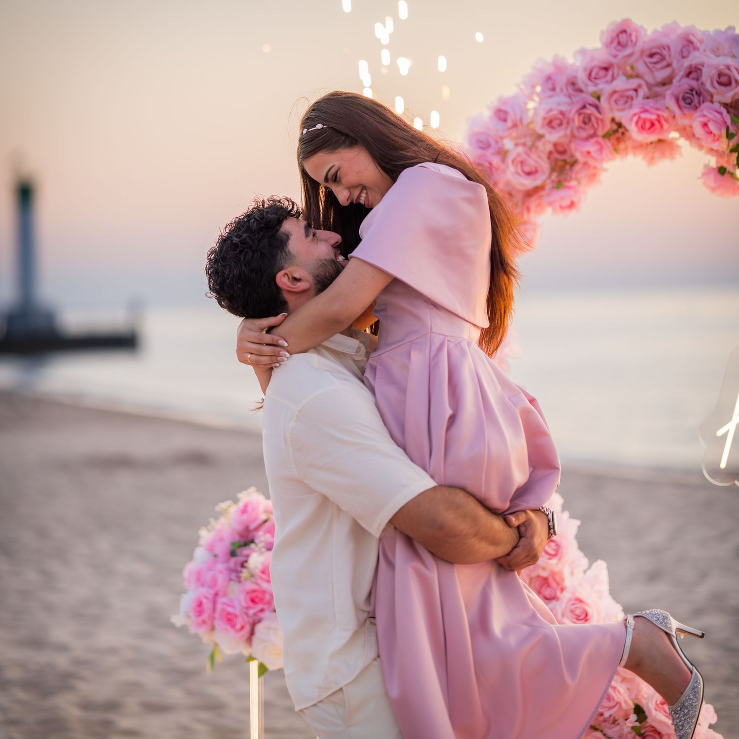 Intimate moment captured during a beach proposal with pink floral decorations and sunset backdrop.