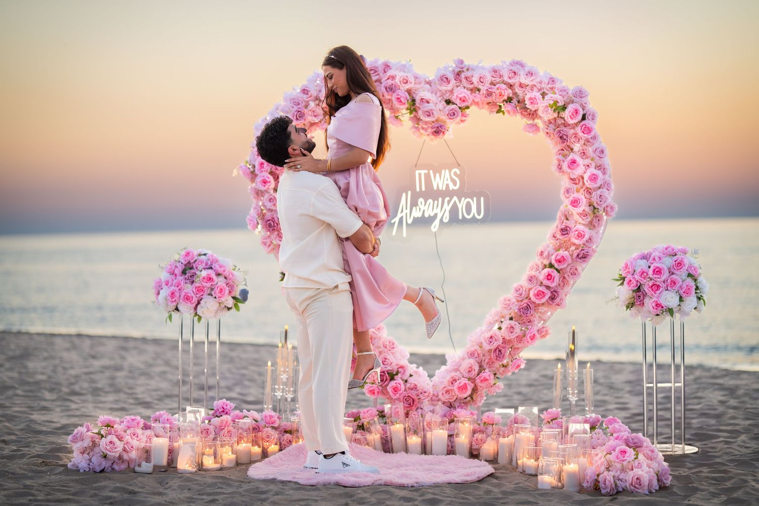 Beach marriage proposal scene with pink floral heart arch and lit candles along the sand.