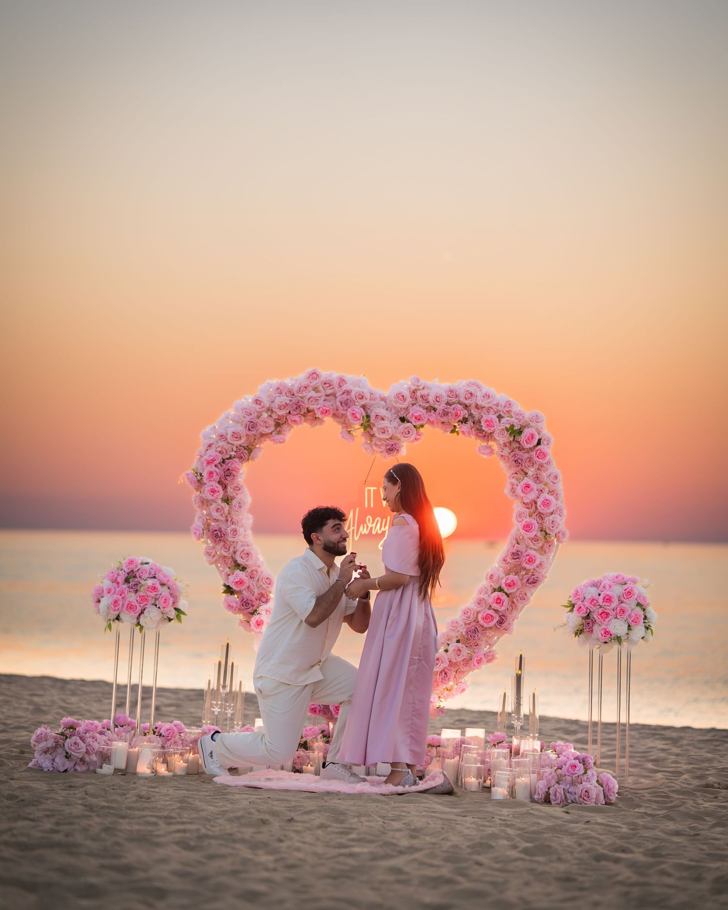 Romantic beach proposal setup with heart-shaped floral arch against sunset sky.