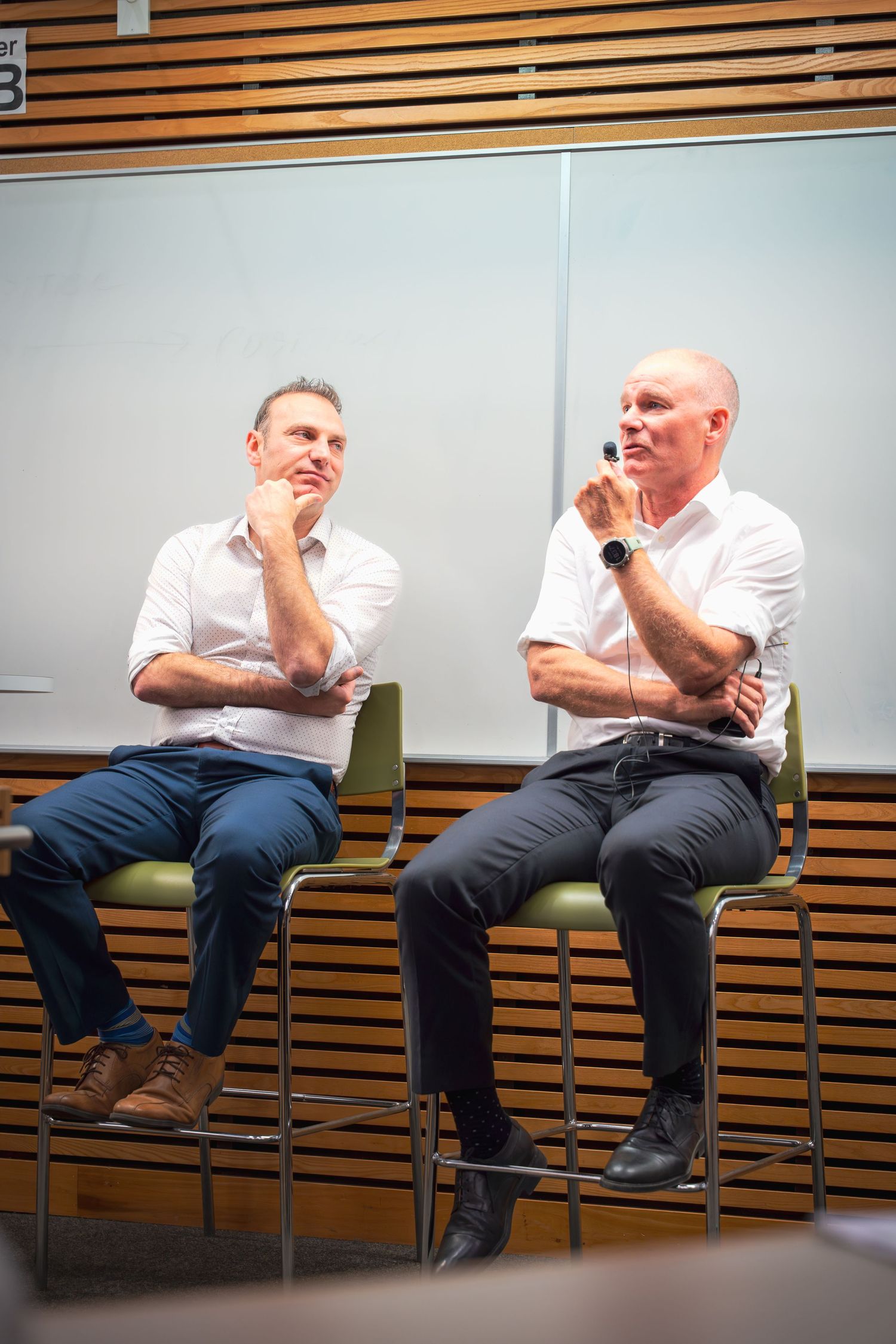 Person in white shirt gesturing while seated in a discussion setting with wooden paneled wall.