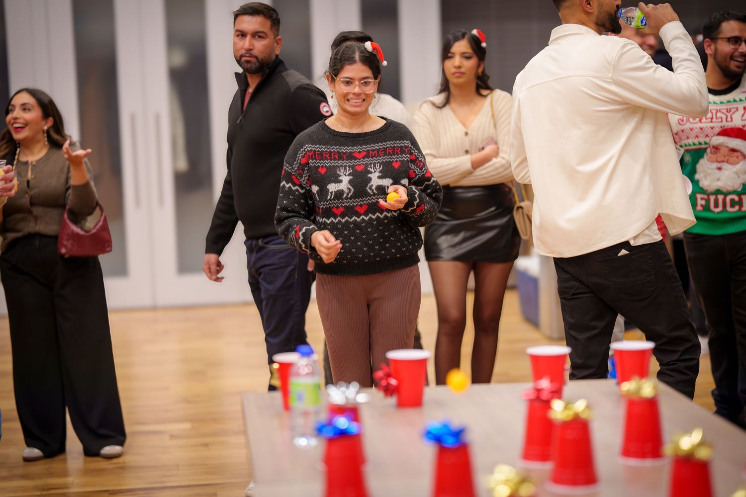 Party guests mingle near tables with red cups in a well-lit room with hardwood flooring.