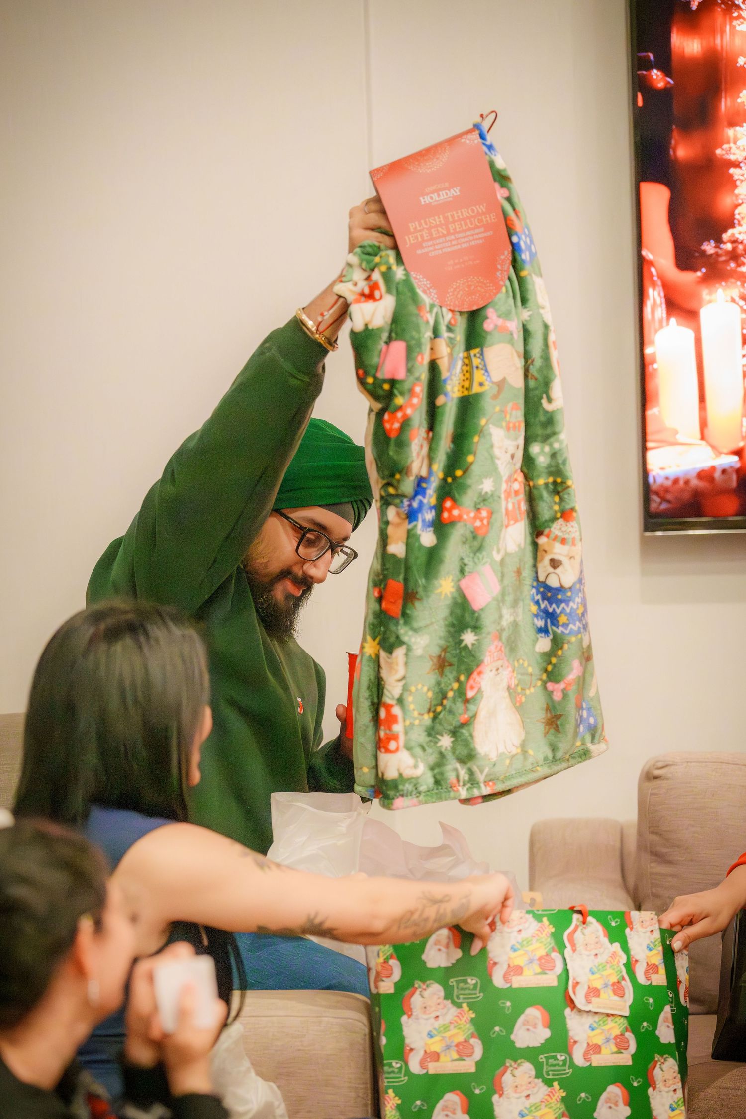 Someone in green clothing unwraps festive holiday-themed gift paper at an indoor gathering.