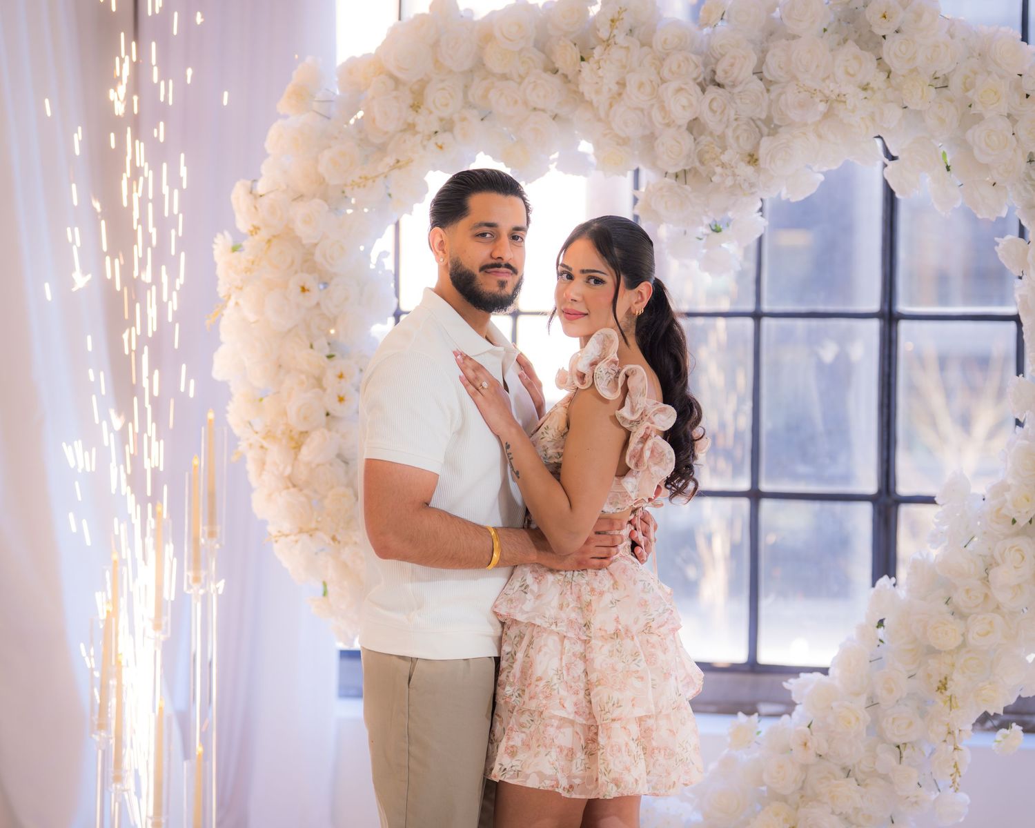Couple embracing inside a heart-shaped white floral arch with sparkling lights.