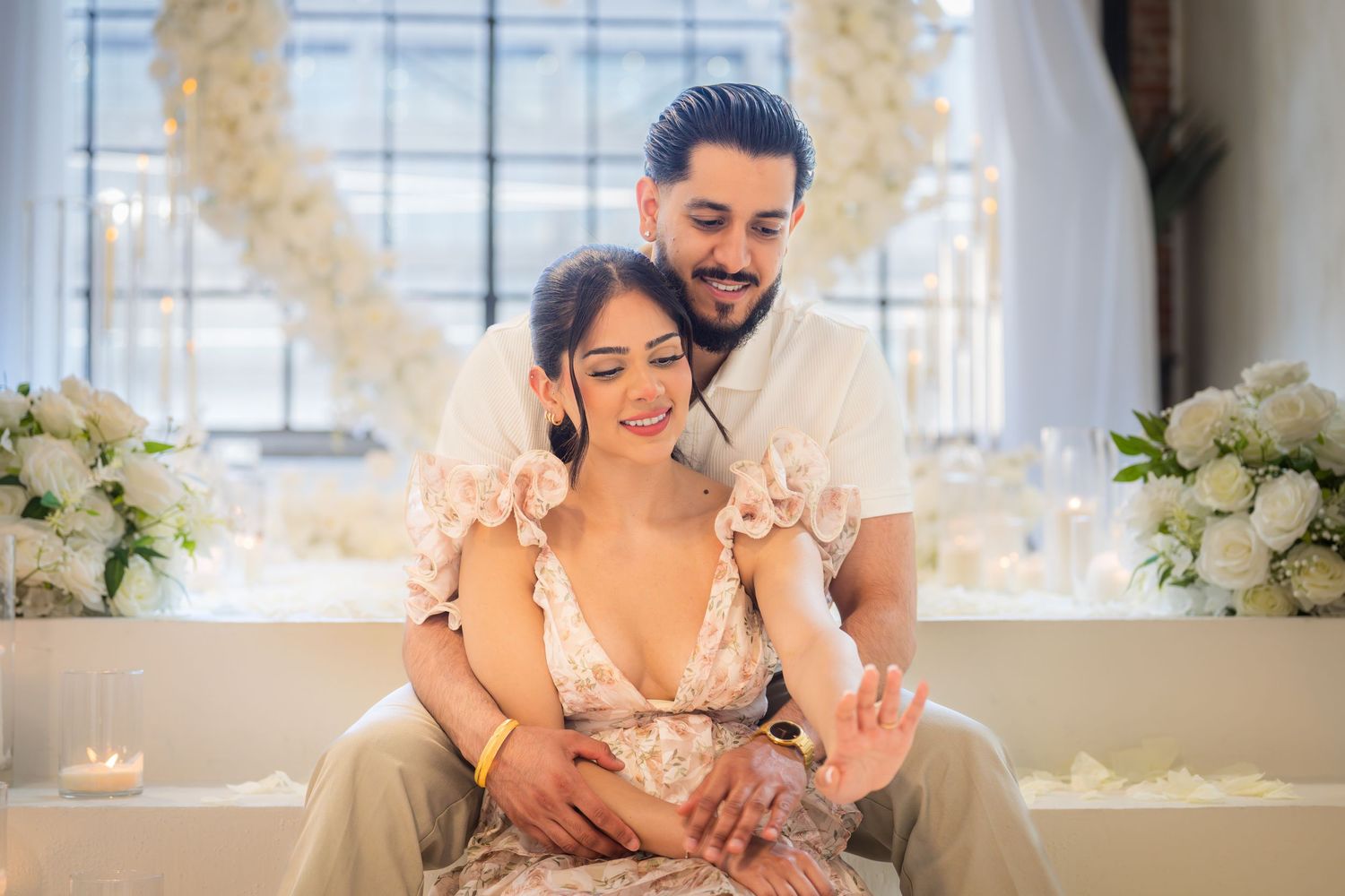 Romantic couple sitting together in front of a white floral heart arch with candles and white flower arrangements.