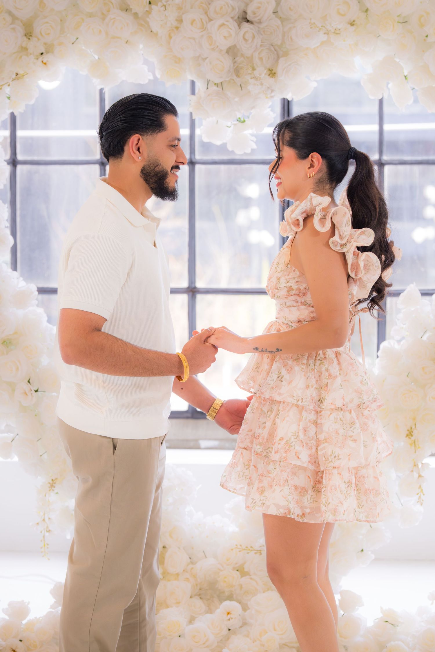 Happy couple holding hands and smiling at each other surrounded by white floral arch backdrop.