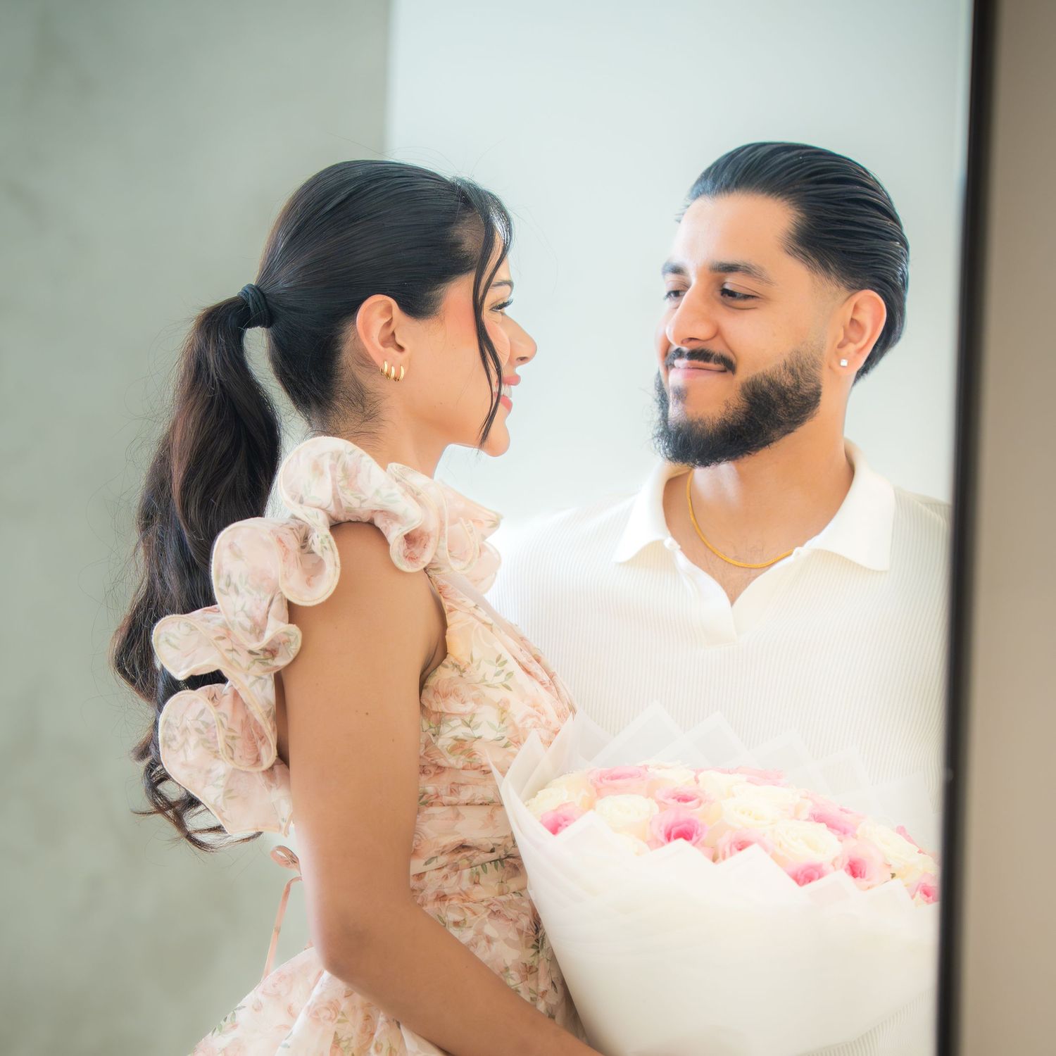 Romantic couple smiling at each other while holding pink and white rose bouquet indoors.