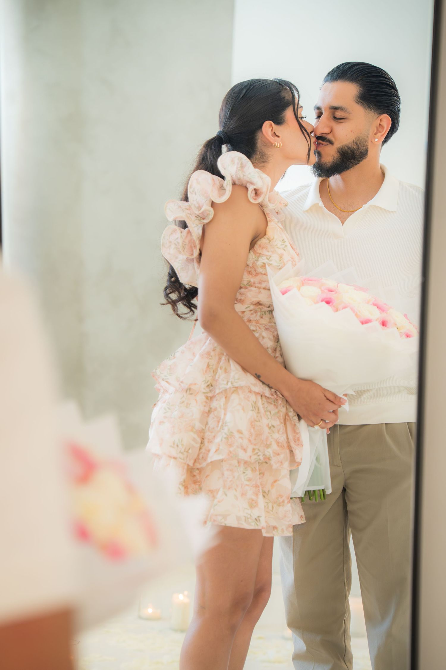 Couple sharing a kiss while holding a bouquet of pink and white roses by a mirror.