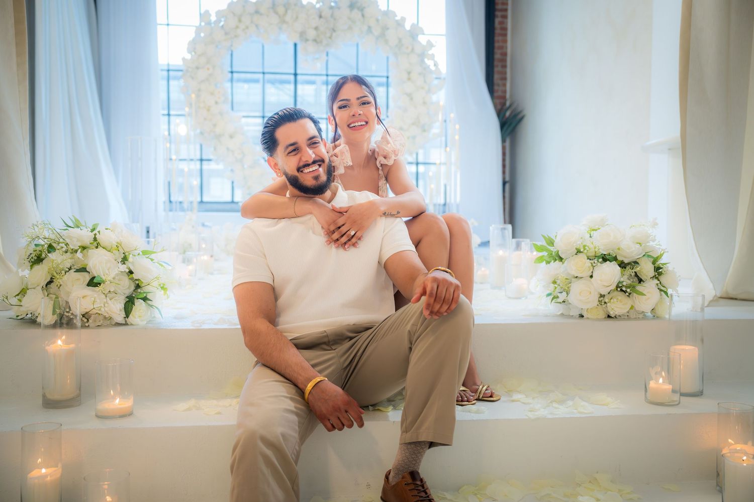 Engaged couple sitting together smiling before heart-shaped white floral arch with candles.