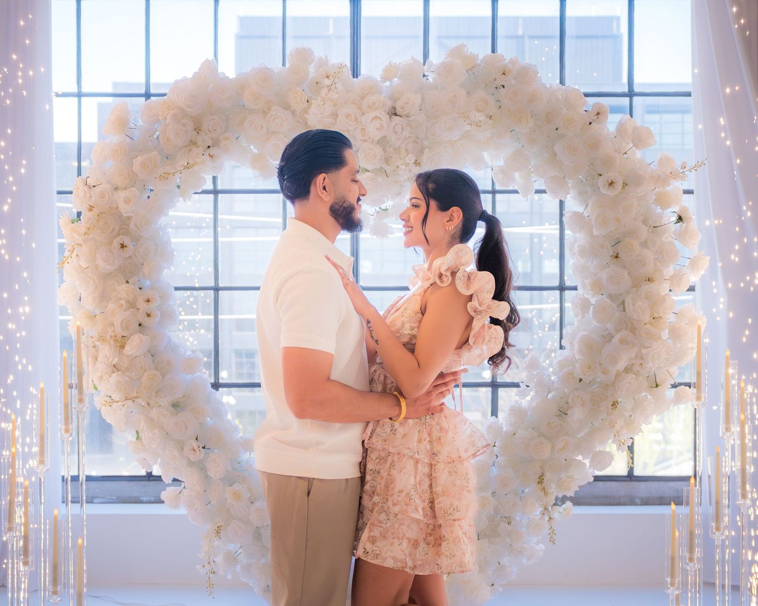 Couple standing together in front of large white floral heart arch during engagement shoot.