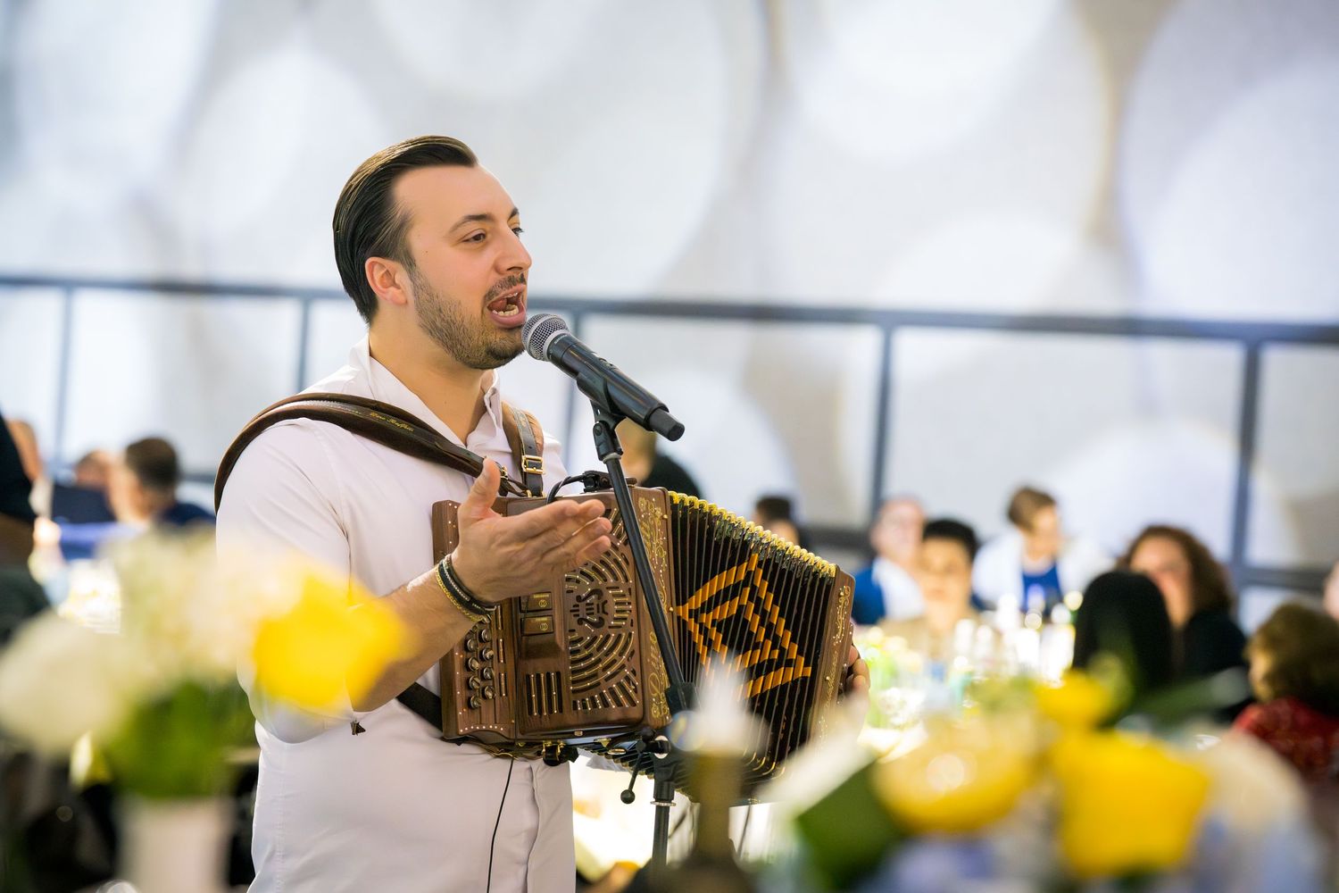 Accordion musician performing and singing at microphone stand during indoor banquet celebration with yellow flowers.