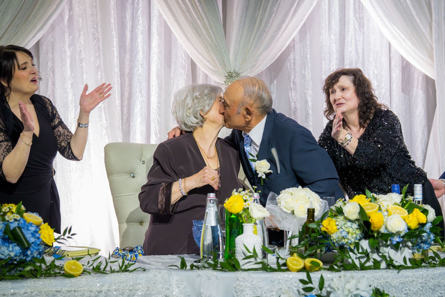 Elderly couple sharing a kiss at a wedding reception table adorned with yellow and blue flowers.