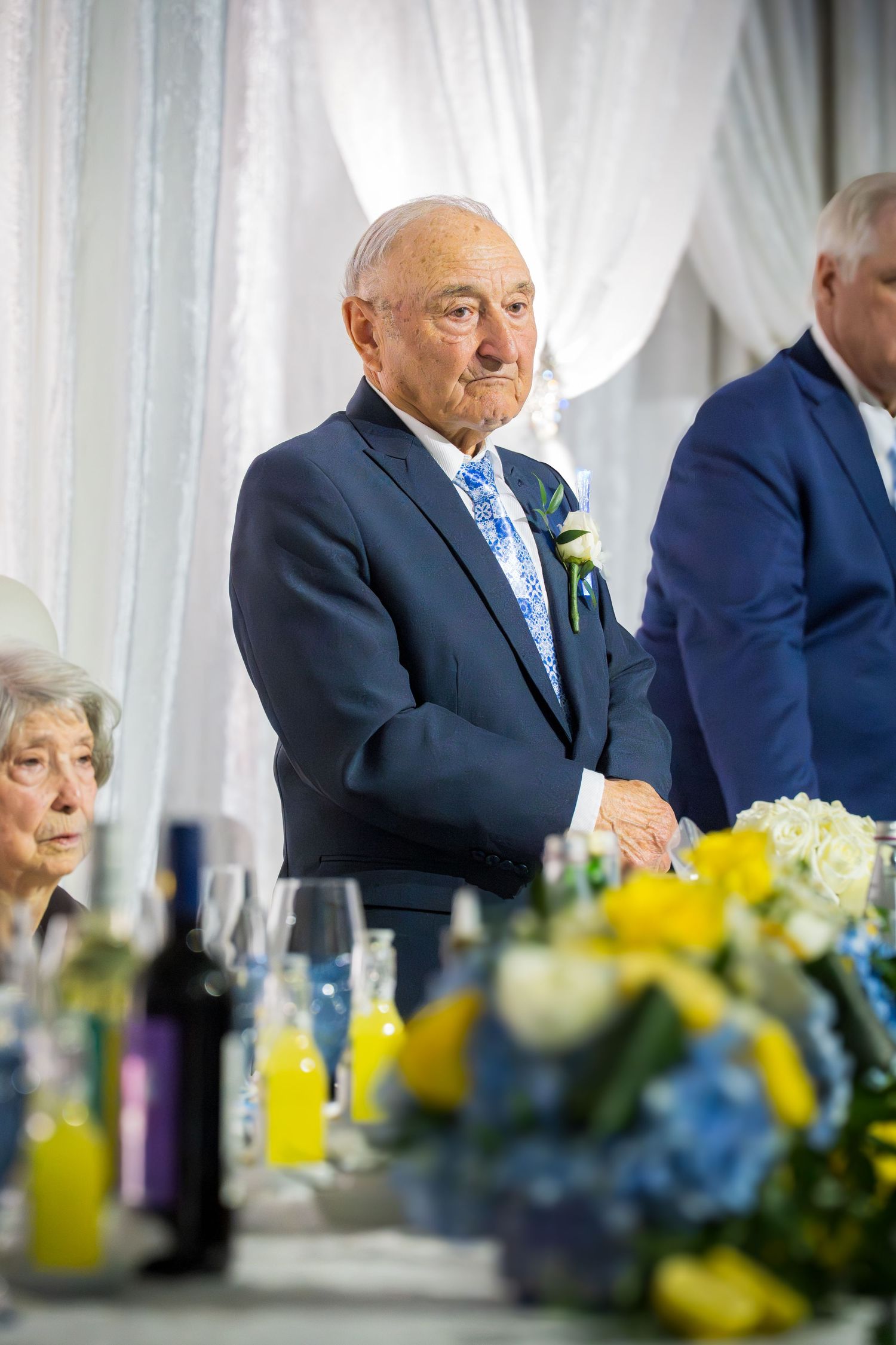 Elderly man in navy suit with floral tie and boutonniere stands at wedding reception table with flowers.