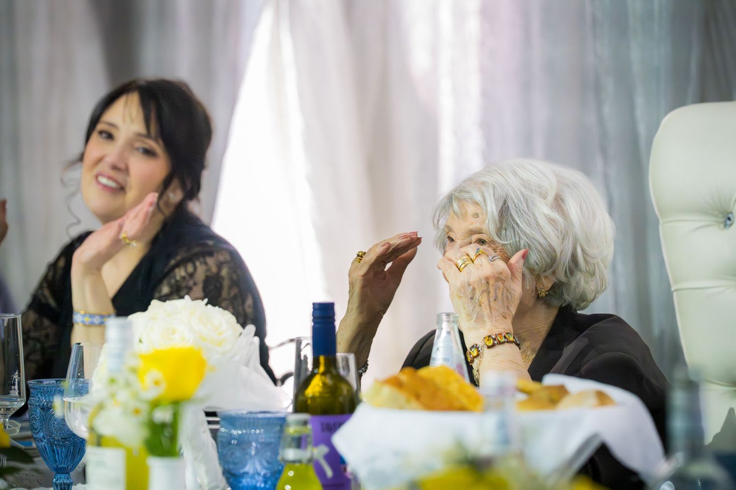 Two women laughing joyfully at a festive dinner table with yellow flowers and wine bottles nearby.