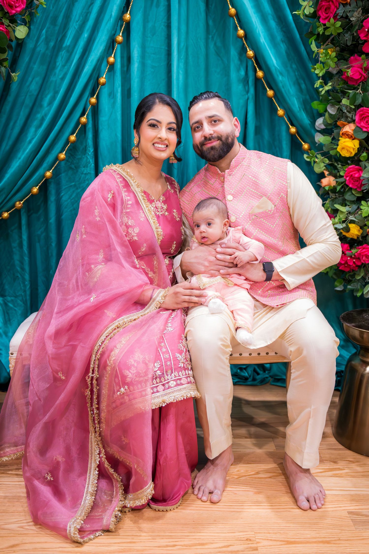 South Asian couple in pink and cream traditional attire seated with newborn at decorated backdrop.