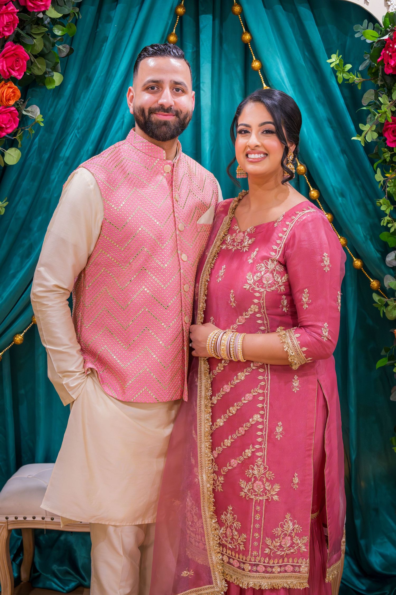 Smiling couple in coordinating pink South Asian traditional attire posing at a decorated event.