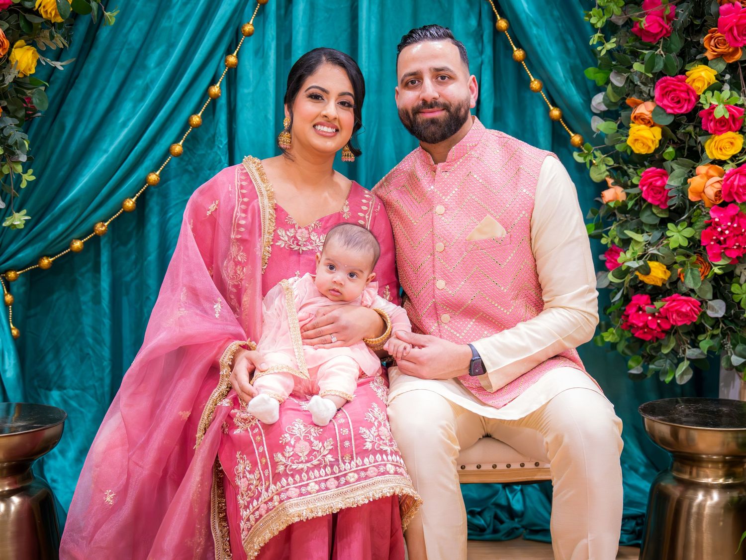 A smiling couple in pink Indian outfits sits together holding their infant baby at a festive celebration.