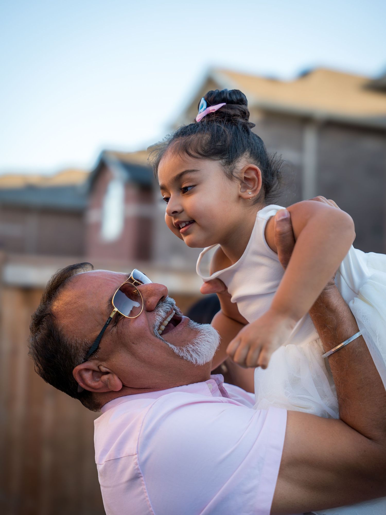 A grandfather in sunglasses joyfully lifts a little girl in a white dress outdoors.