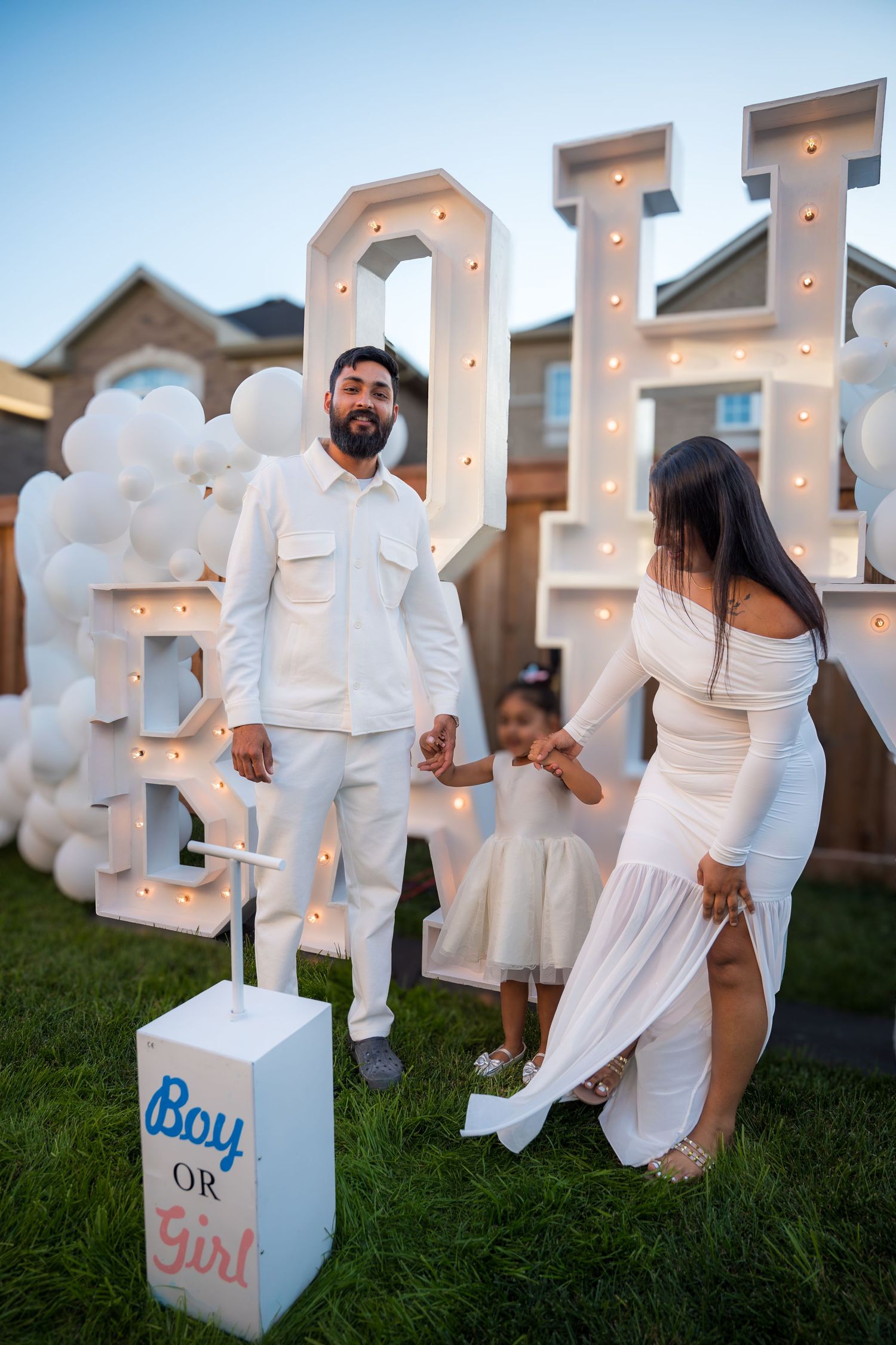 Family in white outfits at a gender reveal party with illuminated OH BABY letters, balloons, and Boy or Girl sign.