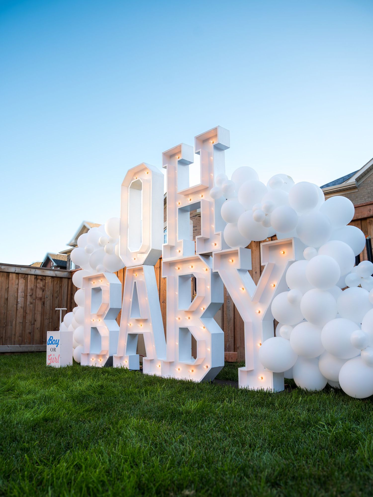 Large illuminated OH BABY marquee letters surrounded by white balloon garland at an outdoor gender reveal party.