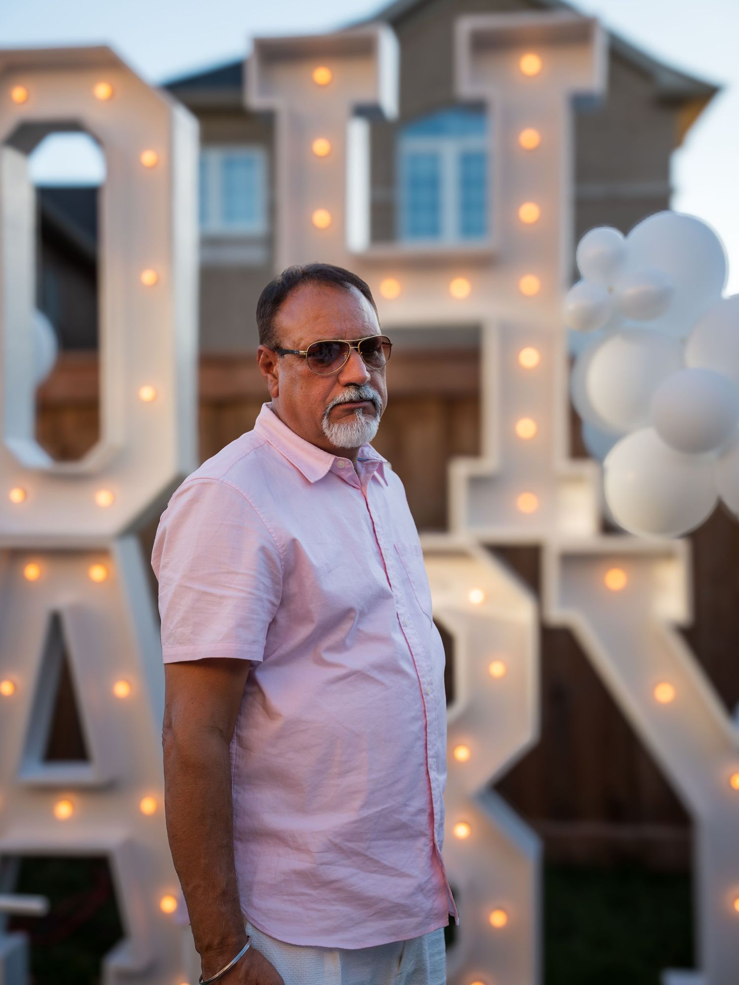 Man in pink shirt and sunglasses poses in front of illuminated OH BABY marquee letters with white balloons at outdoor party.