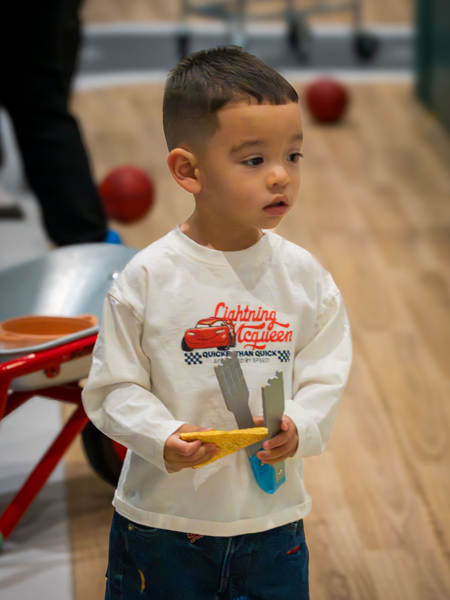 Toddler boy in Lightning McQueen shirt holds a yellow toy while playing at an indoor children's play space.