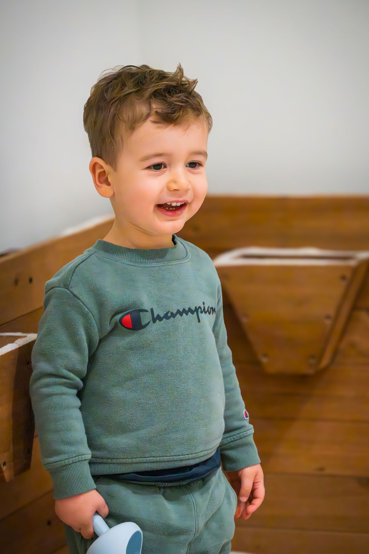 Smiling toddler boy in green Champion sweatshirt playing in a wooden play structure.