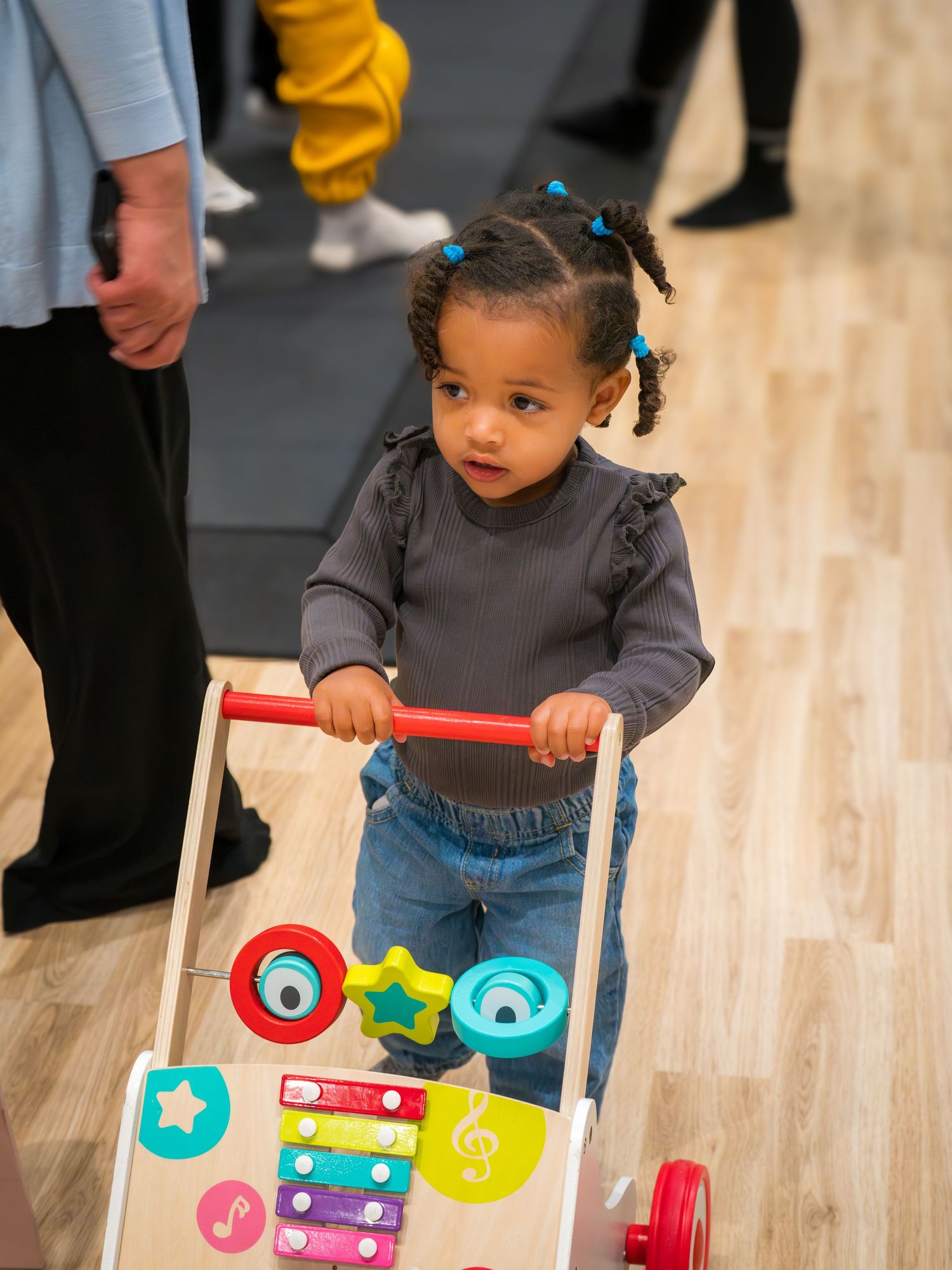 Toddler girl with pigtails pushing a colorful wooden activity walker toy.