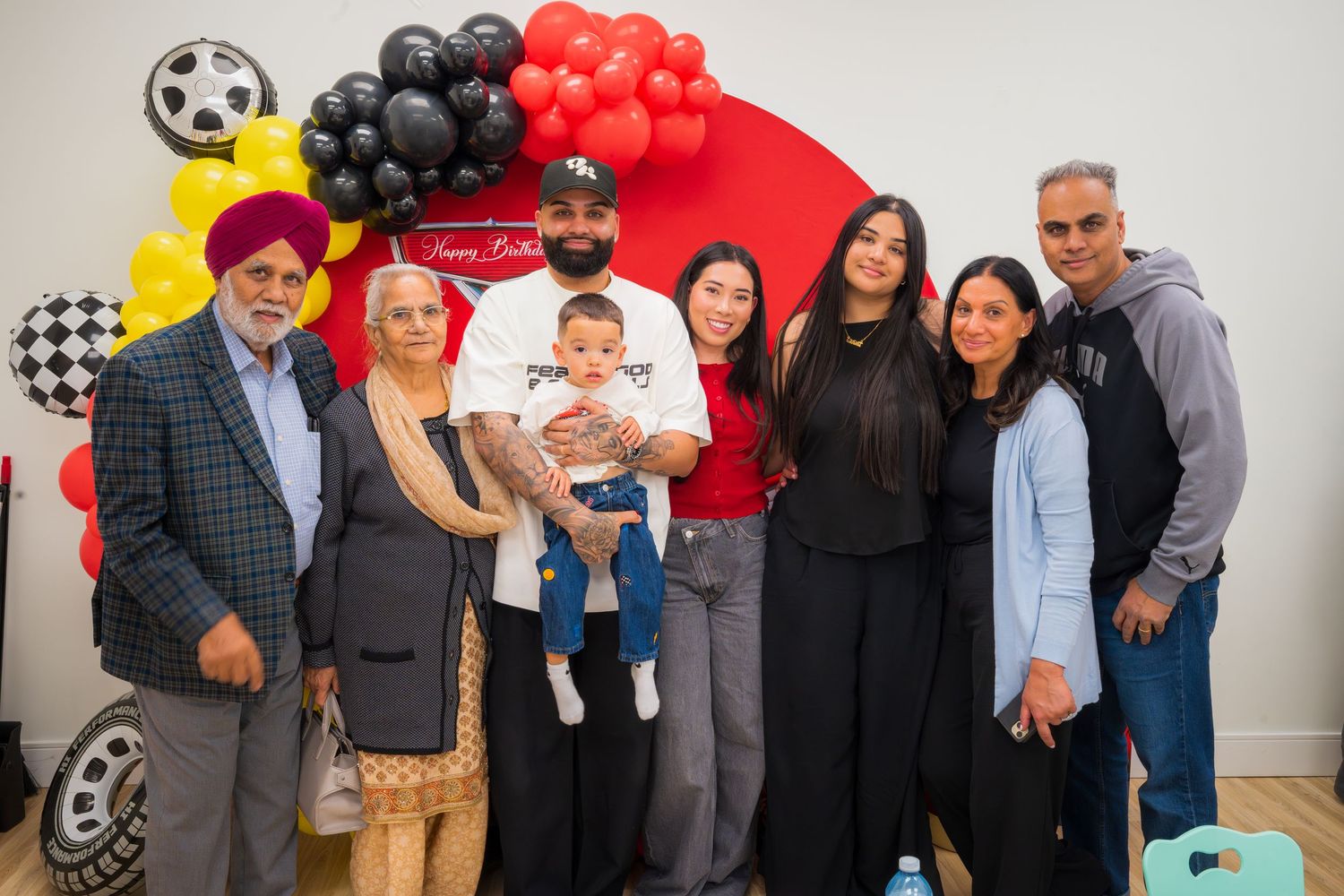 Family group photo at a Cars-themed first birthday party with red and black balloon decorations.