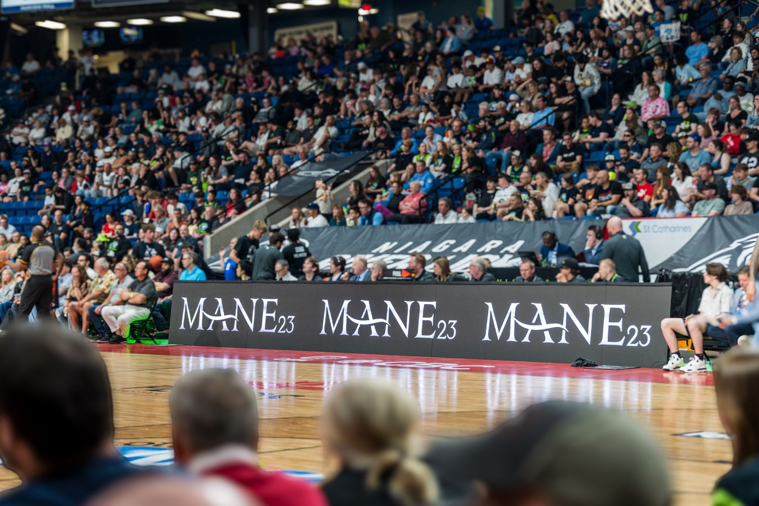 Indoor basketball arena with MANE23 branding displayed on courtside LED boards, surrounded by a packed crowd of spectators.
