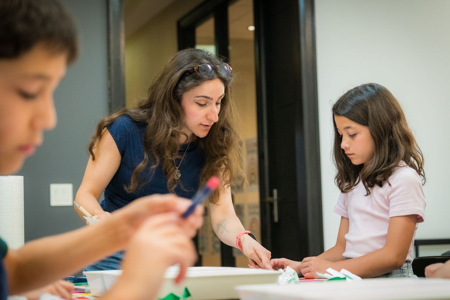 Female teacher assists young girl with colorful paper art project during hands-on children's workshop.
